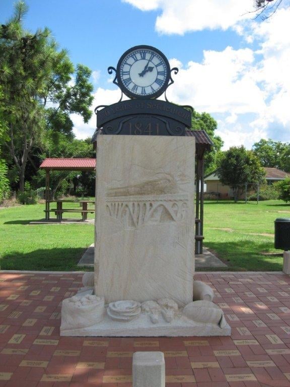 A decorative clock tower with a white stone pedestal base and ornate black clock face, set in a park with green grass and trees