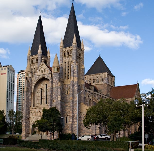 Neo-Gothic stone cathedral with twin spires and black peaked roofs, surrounded by trees and modern buildings