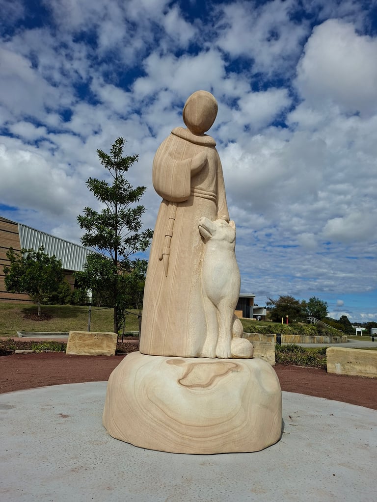 Wooden carved statue of a robed figure holding a child, displayed in an outdoor public space under a blue sky with clouds