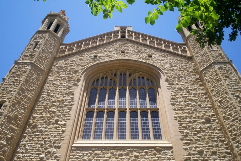 Stone church facade with tall Gothic-style windows and decorative turrets against a blue sky with green foliage visible at edges