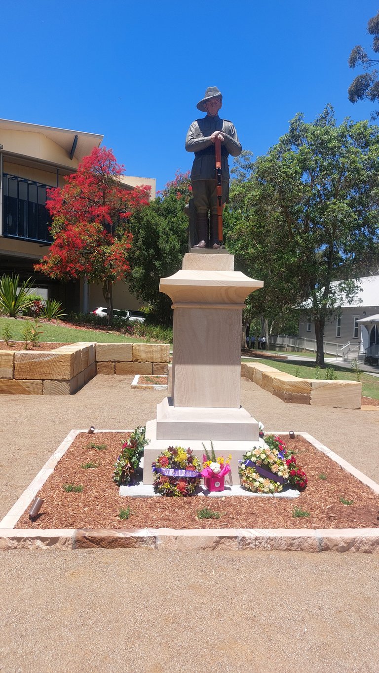 Bronze statue of a man in period clothing standing on a pedestal in a garden setting with colorful flowers and autumn foliage