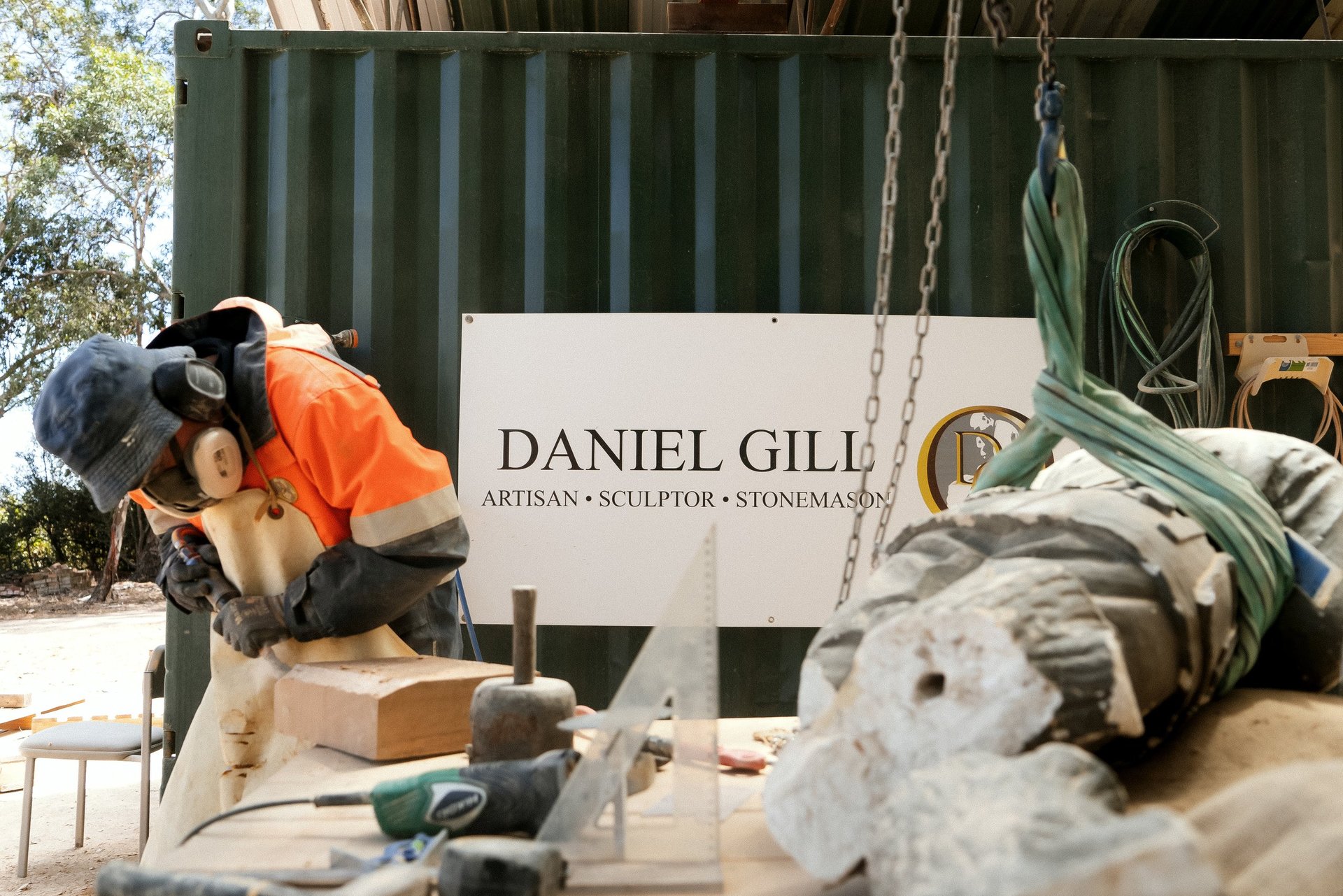 Artisan sculptor working on stone carving in outdoor workshop with sign reading Daniel Gill Artisan Sculptor Stonemason