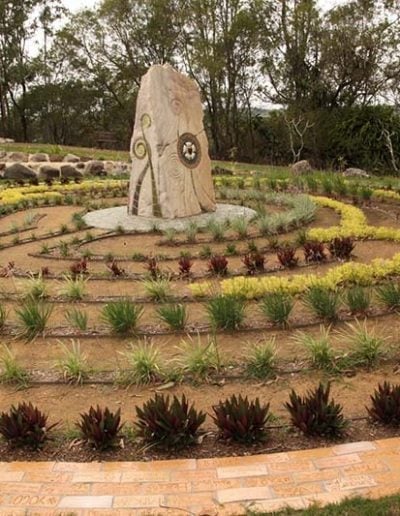 A large stone monument with circular motifs stands at the center of a curved garden bed with layered plantings of native shrubs and grasses