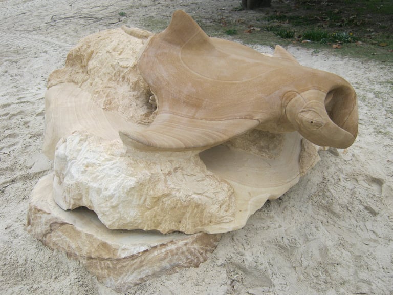 Wooden carving of a fish head in progress, showing natural wood grain and unfinished sculptural details on sandy ground