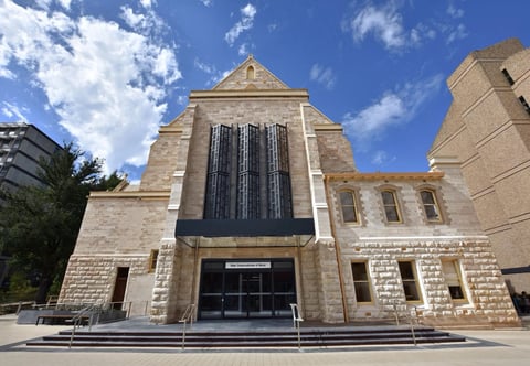 Exterior view of a stone church with contemporary architectural elements, featuring a peaked roof and large dark windows under a blue sky with white clouds.