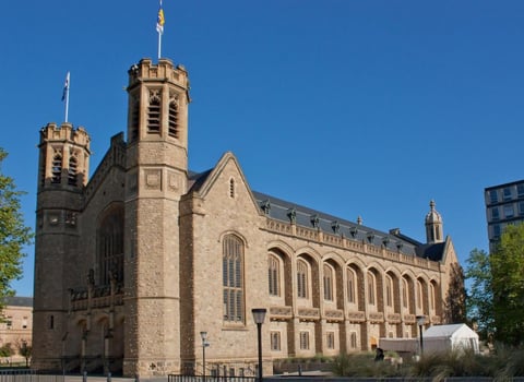 Historic stone church with twin towers and cross spires against blue sky, surrounded by landscaping and buildings