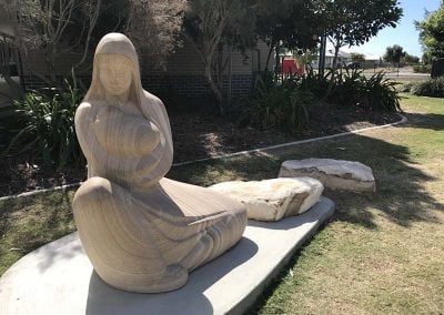 Wooden carved sculpture of a woman sitting in a garden with trees and landscaping in the background