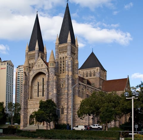 Stone church with twin spires and black peaked roofs, surrounded by green trees, against a blue sky with clouds