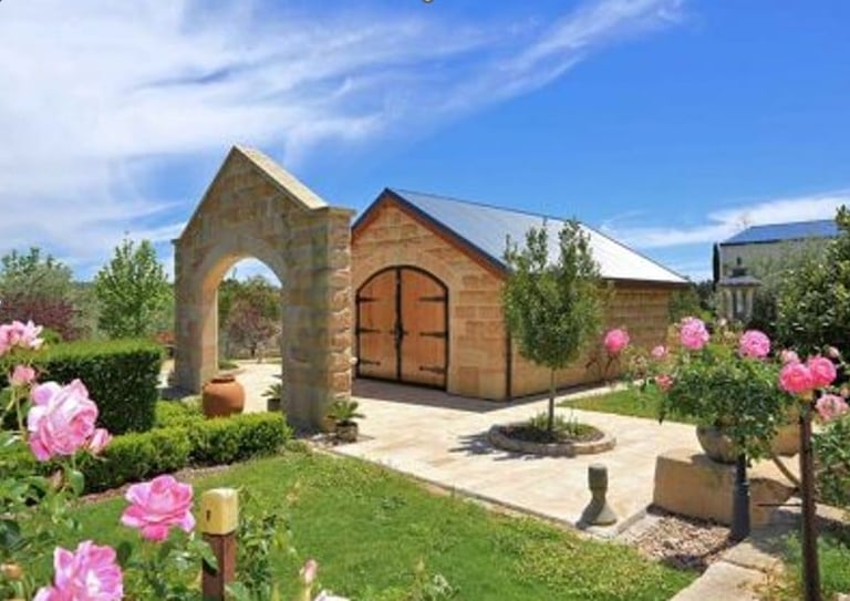 Historic stone building with arched doorway surrounded by pink roses and manicured gardens under blue sky