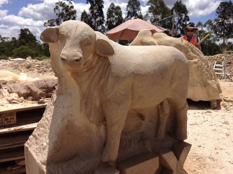 Stone sculpture of a cow in progress at an outdoor quarry or workshop with trees and rocks in background