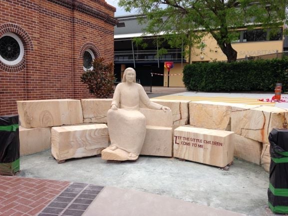 Large wooden carved figure seated on wooden blocks in a plaza with brick buildings in background