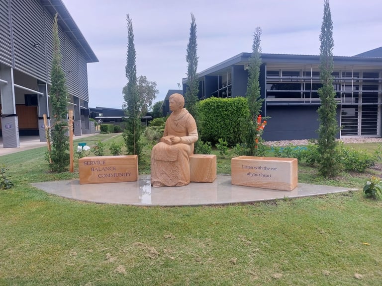 Terracotta sculpture of a seated figure in a courtyard surrounded by green cypress plants and informational plaques, with modern buildings in the background