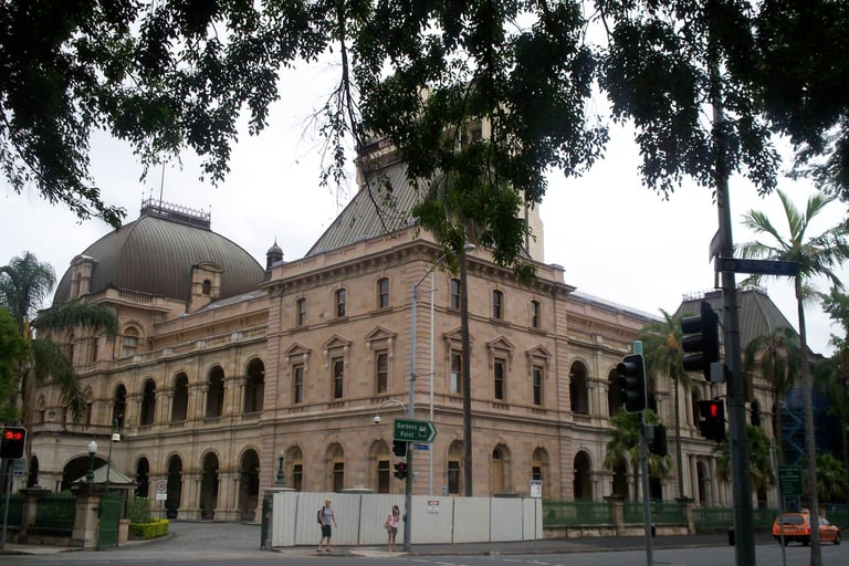 Historic sandstone government building with dome, framed by hanging tree branches and palm trees on street