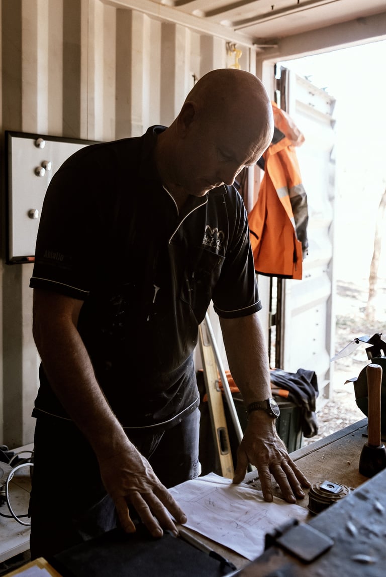 Worker in black shirt leaning over workbench with blueprints and tools, sunlit doorway visible behind