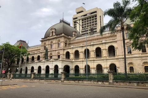 Historic beige stone government building with mansard roof and arched windows, featuring palm trees and modern high-rise building behind